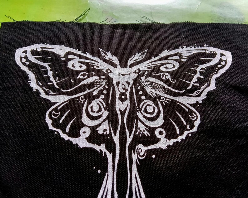 May include: Black fabric patch with a white moth design. The moth's wings and body are intricately detailed. The white design stands out against the black background, creating a bold contrast.