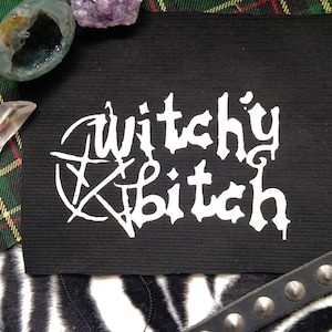 Könnte beinhalten: Weißer Bügel-Patch mit dem Text "Witchy Bitch" in gotischer Schrift. Der Patch befindet sich auf einem schwarzen Stoffhintergrund.