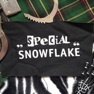 Könnte beinhalten: Schwarzer Patch mit weißem Text, der "Special" in einer ausgeschnittenen Schriftart und "SNOWFLAKE" in einer Blockschriftart lautet. Der Patch ist von einem grün-roten karierten Stoff umgeben.