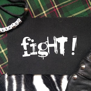 Könnte beinhalten: Ein schwarzes Etikett mit dem Wort "fight!" in weißer, stilisierter Schrift. Das Etikett befindet sich auf einem grün, rot und gelb karierten Stoff. Eine Perlenkette mit den Buchstaben "XXXKYOU" ist sichtbar.