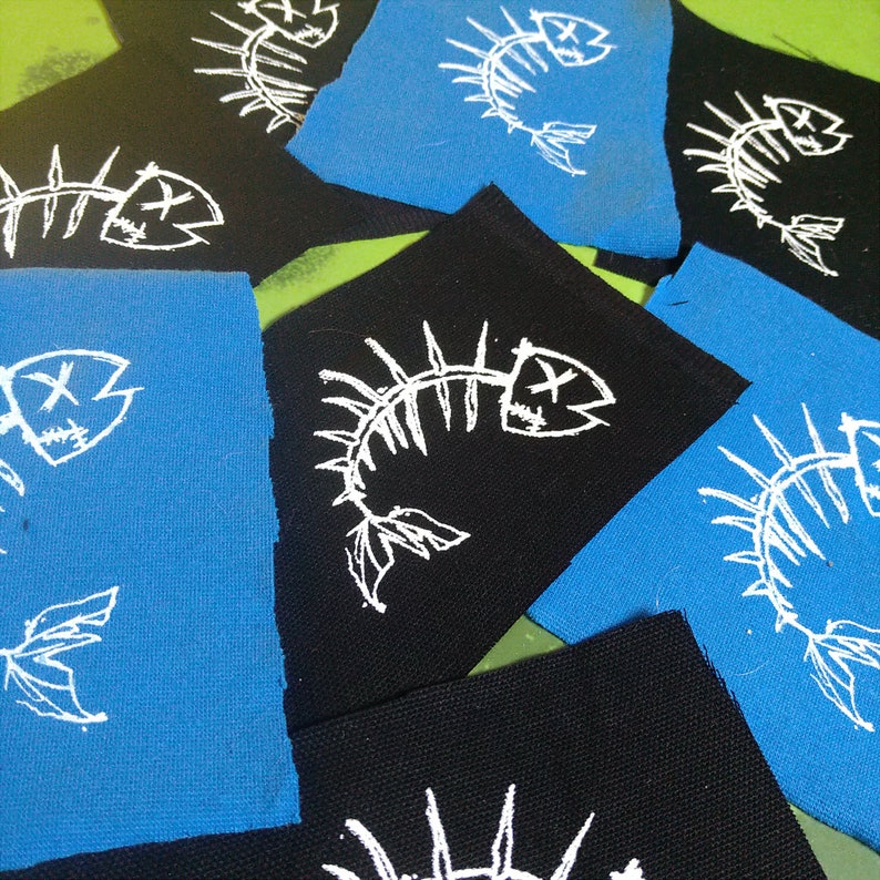 K&ouml;nnte beinhalten: Eine Sammlung von B&uuml;gelbildern mit einem wei&szlig;en Fischskelett-Design auf schwarzem und blauem Stoff.