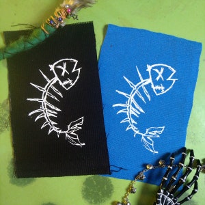 May include: Two iron-on patches featuring a white fish skeleton design on black and blue fabric. The fish skeleton has a skull with an X for eyes.