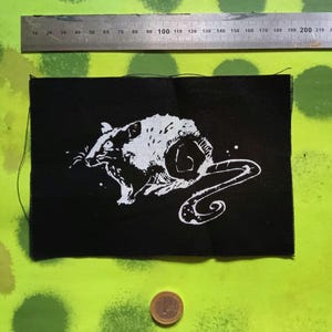 Op de afbeelding: Zwart rechthoekig embleem met een witte illustratie van een rat. De rat wordt afgebeeld in een dynamische pose met een gekrulde staart. Een metalen liniaal met centimeter markeringen staat erboven.