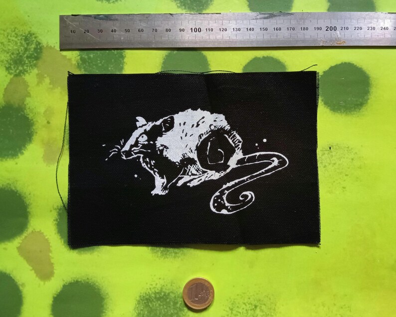 Op de afbeelding: Zwart rechthoekig embleem met een witte illustratie van een rat. De rat wordt afgebeeld in een dynamische pose met een gekrulde staart. Een metalen liniaal met centimeter markeringen staat erboven.