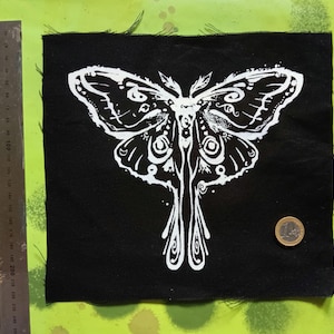 May include: Black square fabric patch featuring a white moth design. The moth has intricate wing patterns and a symmetrical body. The patch has frayed edges. A ruler and a coin are next to the patch.
