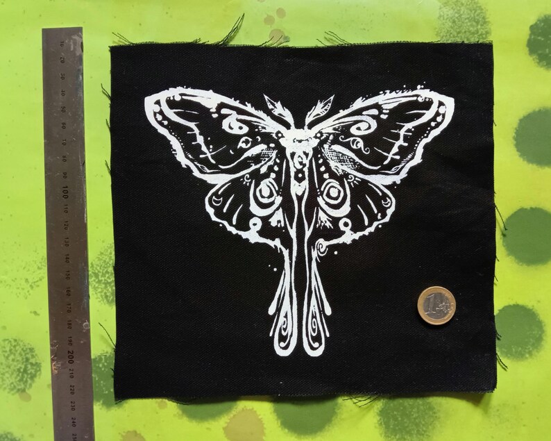 May include: Black square fabric patch featuring a white moth design. The moth has intricate wing patterns and a symmetrical body. The patch has frayed edges. A ruler and a coin are next to the patch.