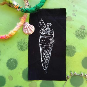 Puede incluir: Un parche de tela negro con un contorno blanco de un cono de helado con una cereza en la parte superior. El cono de helado está goteando helado derretido.
