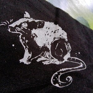 Op de afbeelding: Een zwarte stoffen patch met een witte illustratie van een rat. De rat wordt in profiel getoond, met zijn staart opgerold. Witte spikkels zijn verspreid rond de rat.