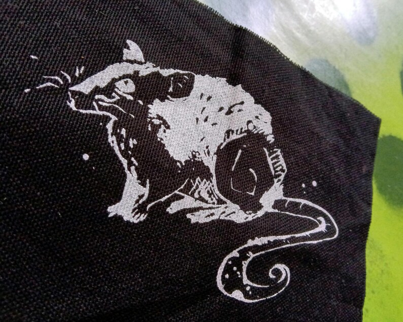Op de afbeelding: Een zwarte stoffen patch met een witte illustratie van een rat. De rat wordt in profiel getoond, met zijn staart opgerold. Witte spikkels zijn verspreid rond de rat.