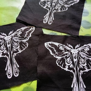 May include: Three black fabric patches with a white moth design. The moth illustration features detailed wing patterns and a stylized body. The patches are arranged on a green surface with white polka dots.
