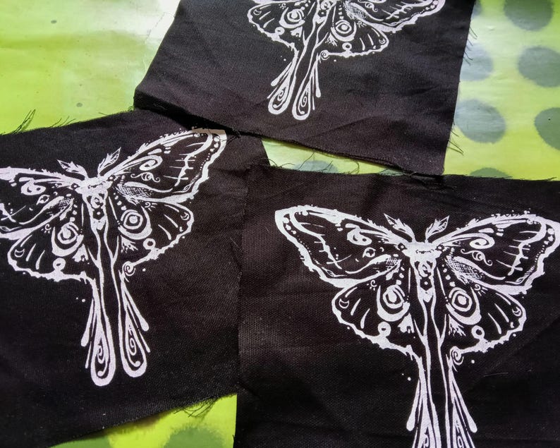 May include: Three black fabric patches with a white moth design. The moth illustration features detailed wing patterns and a stylized body. The patches are arranged on a green surface with white polka dots.