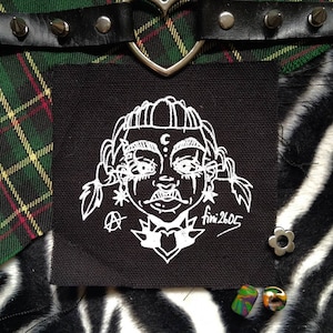 May include: A black square patch with a white illustration of a face with a crescent moon, stars, and the text "fini2605". The patch is on a green and yellow plaid fabric, with a black studded collar and a zebra print background.