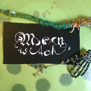 Könnte beinhalten: Schwarzer Stoff-Patch mit weißer Aufschrift "Moon Witch" und einem stilisierten Mond- und Erd-Motiv.