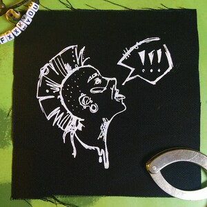 Peut inclure: Patch brodé blanc sur tissu noir représentant un personnage punk avec une crête iroquoise et une bulle de dialogue avec des points d'exclamation.