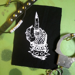 Könnte beinhalten: Schwarz-weiß gestickter Patch einer Hand, die den Mittelfinger zeigt, mit einem Messer im Mittelfinger. Die Hand trägt ein Armband mit Spikes. Der Patch befindet sich auf einem schwarzen Stoffhintergrund.