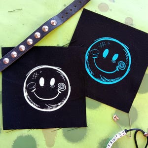May include: Two black fabric patches with white and blue smiley faces printed on them. The smiley faces have a swirl design in the eyes and a line for a mouth.