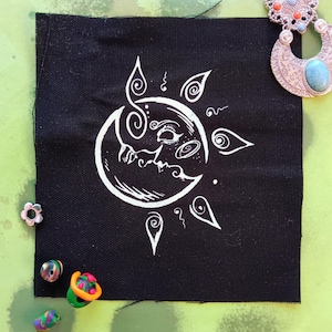 Könnte beinhalten: Ein schwarzer Stoff-Patch mit einem weißen Design einer Sonne und eines Mondes mit einem Gesicht. Sonne und Mond sind ineinander verschlungen und haben ein wirbelndes Design.