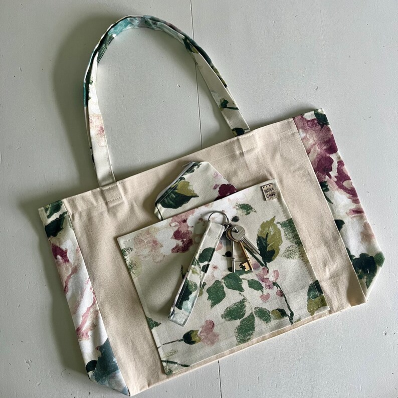 May include: A beige canvas tote bag with floral print fabric panels. The bag has a large pocket on the front with a smaller pocket inside. A key chain with keys is hanging from the pocket.