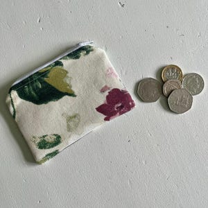 May include: A small white zippered pouch with a floral print featuring pink, green, and yellow flowers. The pouch is lying on a white surface next to a pile of British coins.