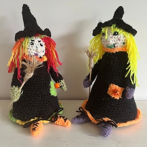 May include: Two handmade crochet witch dolls. Each doll wears a black dress, a pointed black hat, and has yarn hair. One has red and yellow hair, the other yellow. Both hold a broom and have a pumpkin detail.