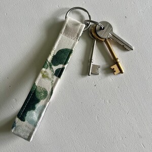 May include: A white fabric key chain with a floral pattern in shades of green. The key chain has a silver key ring with three keys attached.