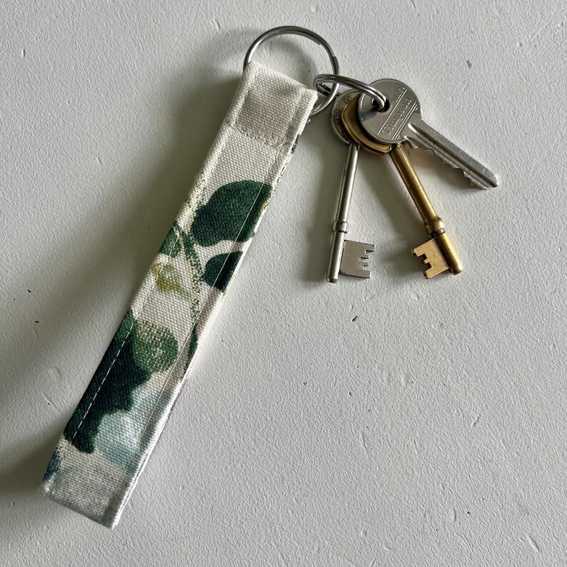 May include: A white fabric key chain with a floral pattern in shades of green. The key chain has a silver key ring with three keys attached.