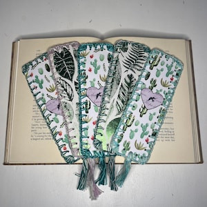May include: Five handmade bookmarks with a white background and a green and white floral pattern. The bookmarks have a heart-shaped design with a bird and text that reads "Hope is the only cure". The bookmarks have a crocheted border and a tassel.