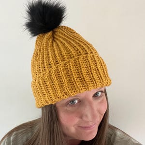 May include: A mustard yellow crocheted beanie with a black faux fur pom pom.
