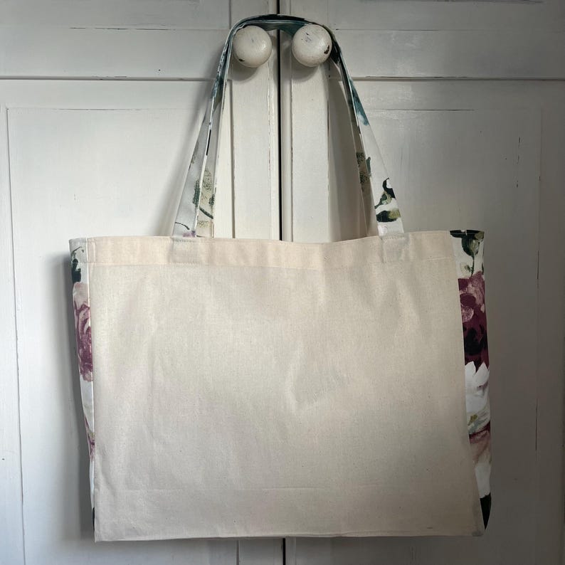 May include: A large, white canvas tote bag with floral print fabric on the sides and handles. The bag has a wide, flat base and is ideal for carrying groceries or other items.