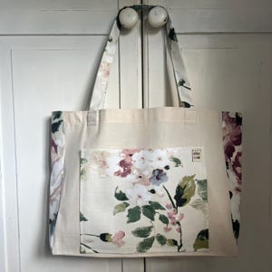 May include: A beige tote bag with a floral print pocket. The bag has two handles and a cream background with a floral print on the sides. The pocket has a floral print with pink, white, and green colours. The text "Little Crafty" is printed on the pocket.