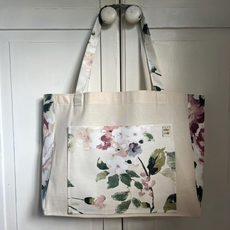 May include: A beige tote bag with a floral print pocket. The bag has two handles and a cream background with a floral print on the sides. The pocket has a floral print with pink, white, and green colours. The text "Little Crafty" is printed on the pocket.