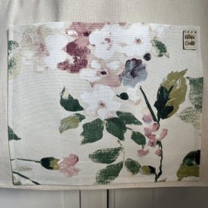 May include: A white cotton apron with a floral print pocket. The pocket has a pink, white, and green floral pattern. The apron has a small brown label with the text "Katona Crafts" on it.