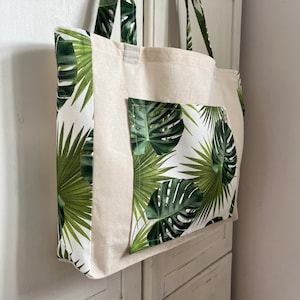 May include: A beige canvas tote bag with a tropical leaf print. The bag has a large front pocket and side panels with a white background and green palm and monstera leaves. The handles and top trim are beige.