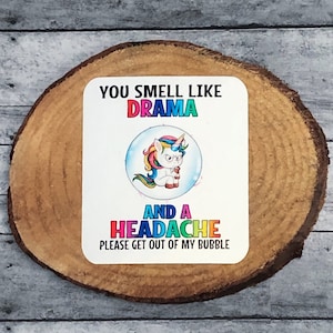 May include: A white coaster with a colorful unicorn inside a bubble. The text reads "You smell like drama and a headache. Please get out of my bubble."