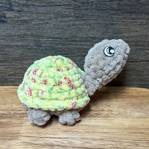May include: A crocheted stuffed turtle toy with a green and white shell with red flowers and a gray body. The turtle has a black eye.