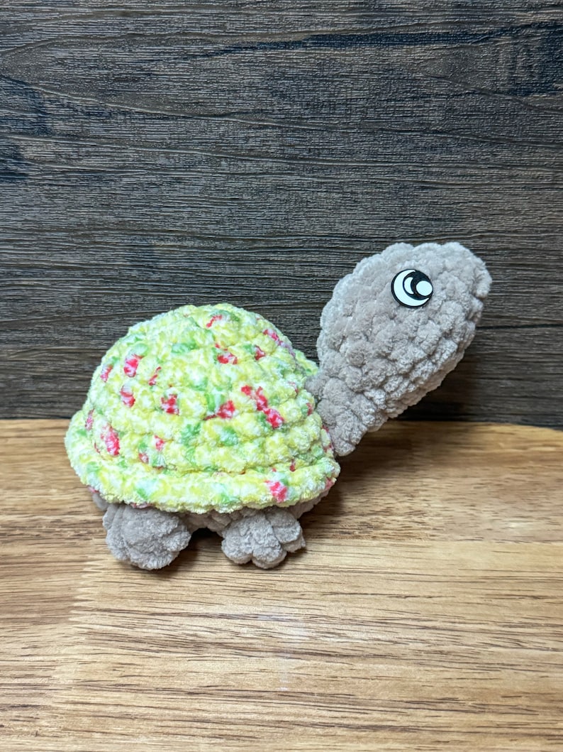 May include: A crocheted stuffed turtle toy with a green and white shell with red flowers and a gray body. The turtle has a black eye.