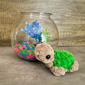 May include: A handmade plush turtle with a green shell and tan body sits next to a glass fishbowl filled with colorful gravel and blue artificial plants. The turtle has a cute, round shape and a friendly appearance.