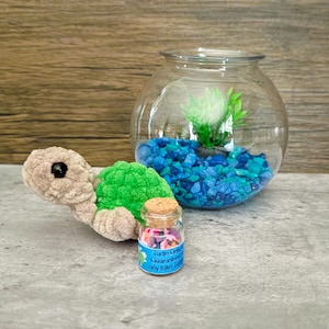 May include: A plush turtle toy with a green shell and tan body sits next to a glass fishbowl filled with blue stones and a small plant. A tiny glass jar with a cork stopper and colorful contents is also present.