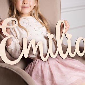 Op de afbeelding: Houten bord met de naam "Emilia" in een cursief lettertype, met hartaccenten boven de letters "i". Het bord is gemaakt van lichtgekleurd hout en wordt vastgehouden door een persoon die een witte top en een roze rok draagt.