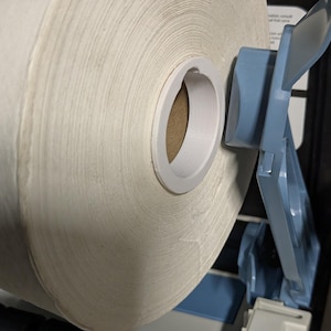May include: A large roll of white paper towels mounted on a blue and white dispenser. The paper towels are tightly wound, and the dispenser has a tear bar for easy access. The dispenser is designed for commercial or industrial use.