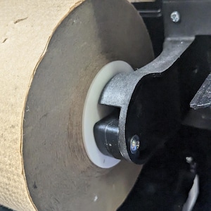 May include: Close-up of a large roll of brown paper towel in a dispenser. The paper towel roll is mounted on a white plastic ring and held in place by a black metal arm. The dispenser is black and has a level on top.