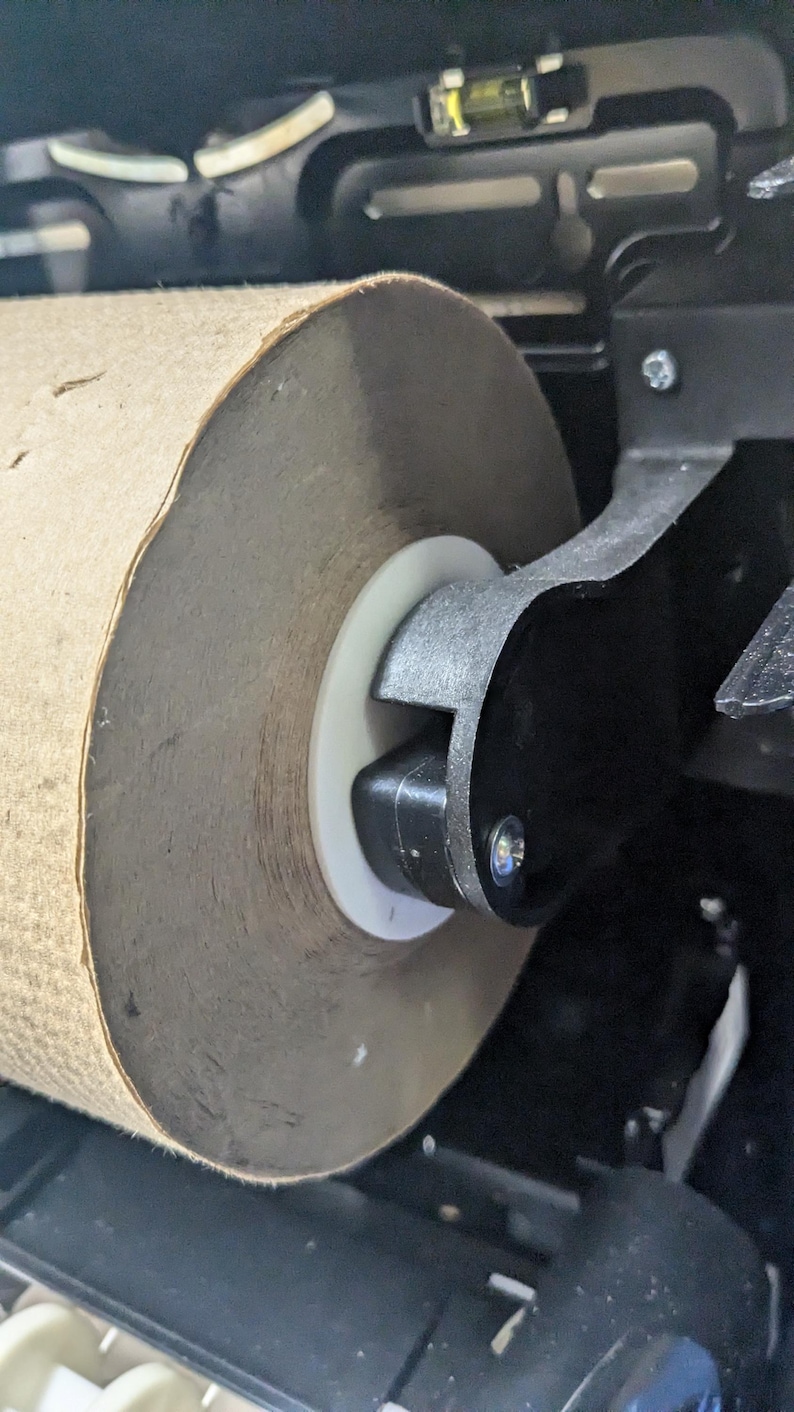 May include: Close-up of a large roll of brown paper towel in a dispenser. The paper towel roll is mounted on a white plastic ring and held in place by a black metal arm. The dispenser is black and has a level on top.