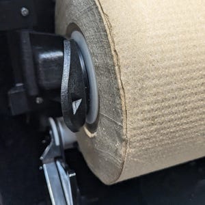 May include: Close-up of a paper towel dispenser. A large roll of brown paper towels is mounted inside a black dispenser. A black lever is visible on the side, and the dispenser is partially open.