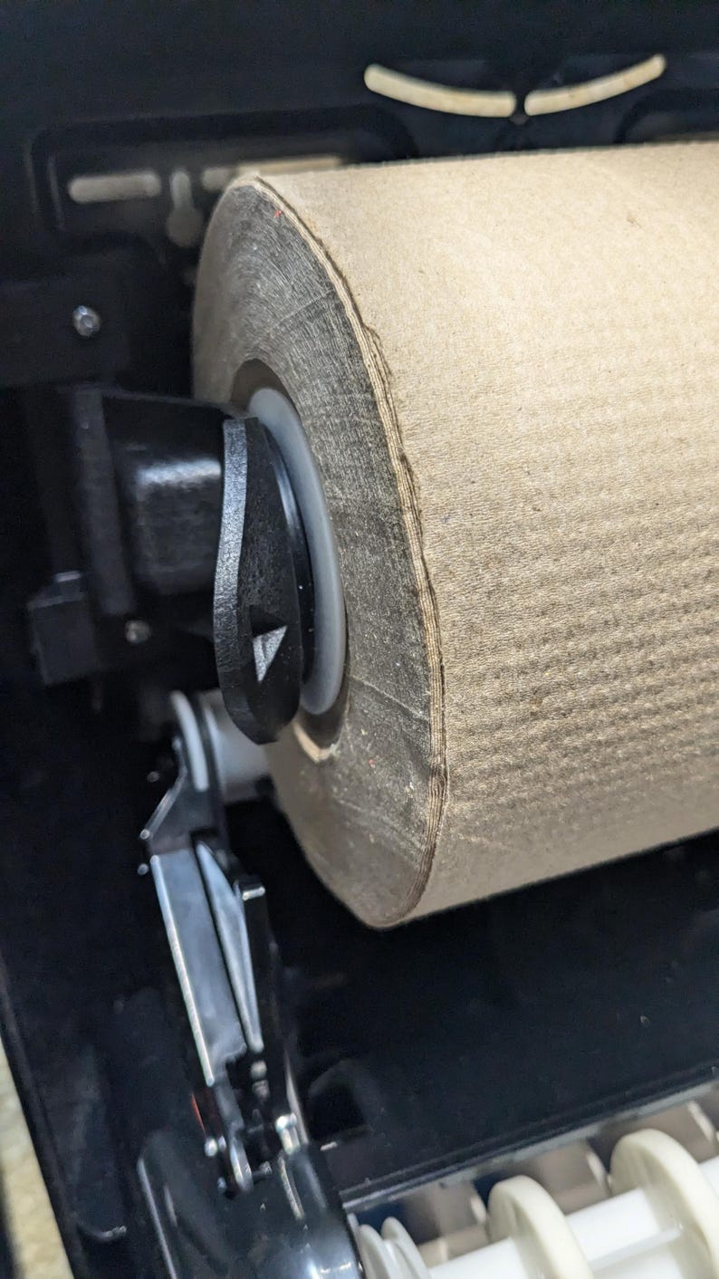 May include: Close-up of a paper towel dispenser. A large roll of brown paper towels is mounted inside a black dispenser. A black lever is visible on the side, and the dispenser is partially open.