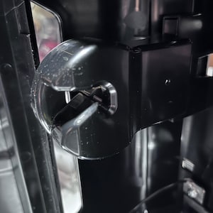 May include: Close-up of a black plastic component with a clear, curved section. The component has a metallic pin and is part of a larger, dark-colored machine. The design features sharp angles and a glossy finish.