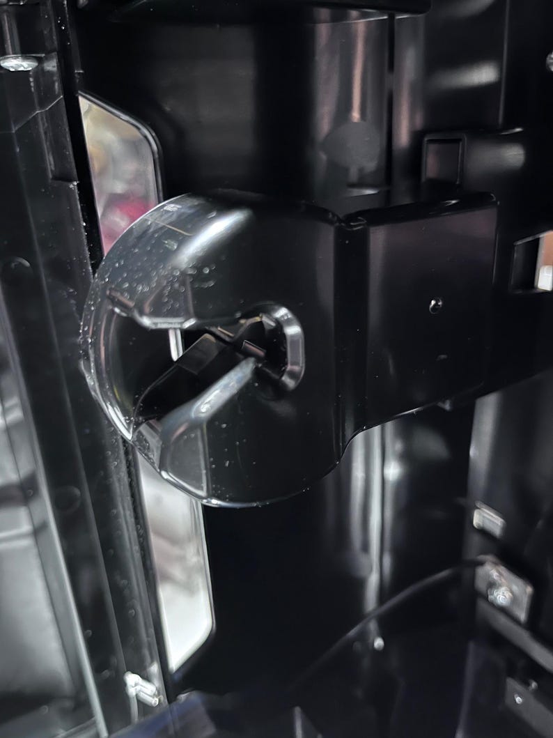 May include: Close-up of a black plastic component with a clear, curved section. The component has a metallic pin and is part of a larger, dark-colored machine. The design features sharp angles and a glossy finish.