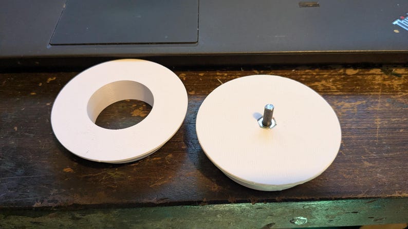 May include: Two white, circular components. One is a solid disc with a metal rod protruding from the center. The other is a white ring. Both are on a dark wooden surface, likely parts of a mechanical assembly.