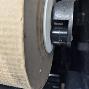 May include: Close-up of a large roll of brown paper towel in a dispenser. The paper roll is mounted on a white plastic disc and held in place by a black plastic mechanism. The dispenser is black and white.