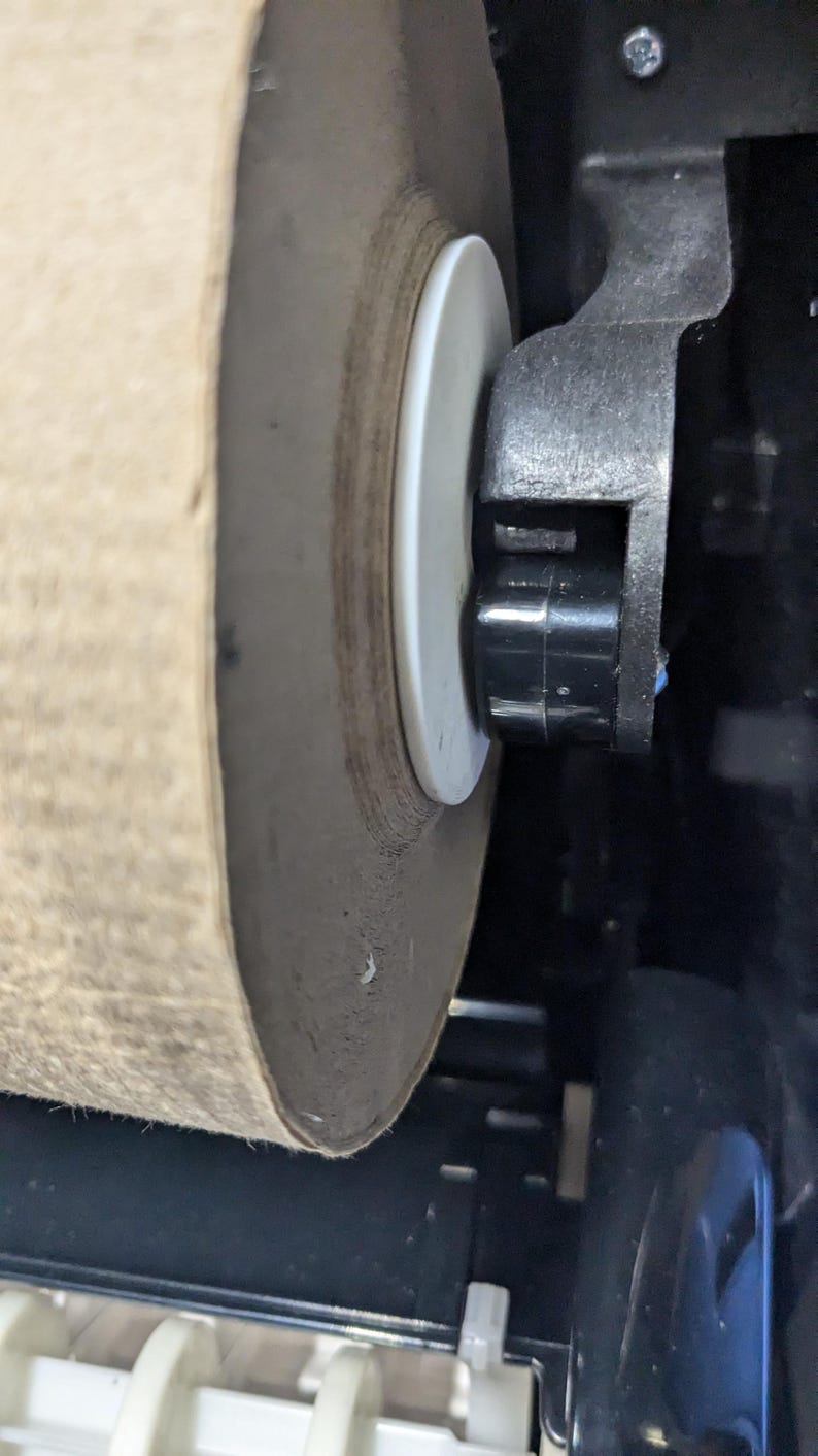 May include: Close-up of a large roll of brown paper towel in a dispenser. The paper roll is mounted on a white plastic disc and held in place by a black plastic mechanism. The dispenser is black and white.
