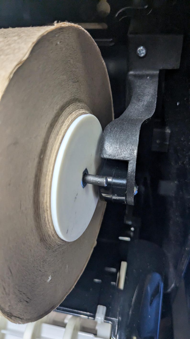 May include: Close-up of a large roll of brown paper towels inside a black dispenser. The roll is mounted on a white plastic disc and a metal rod. The dispenser's black arm holds the roll in place.